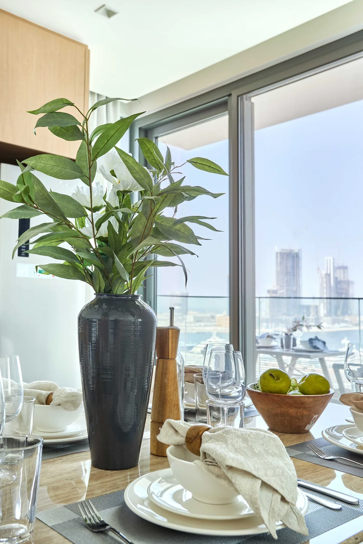 A dining table is set beside a large window, with a leafy plant and fruit bowl.