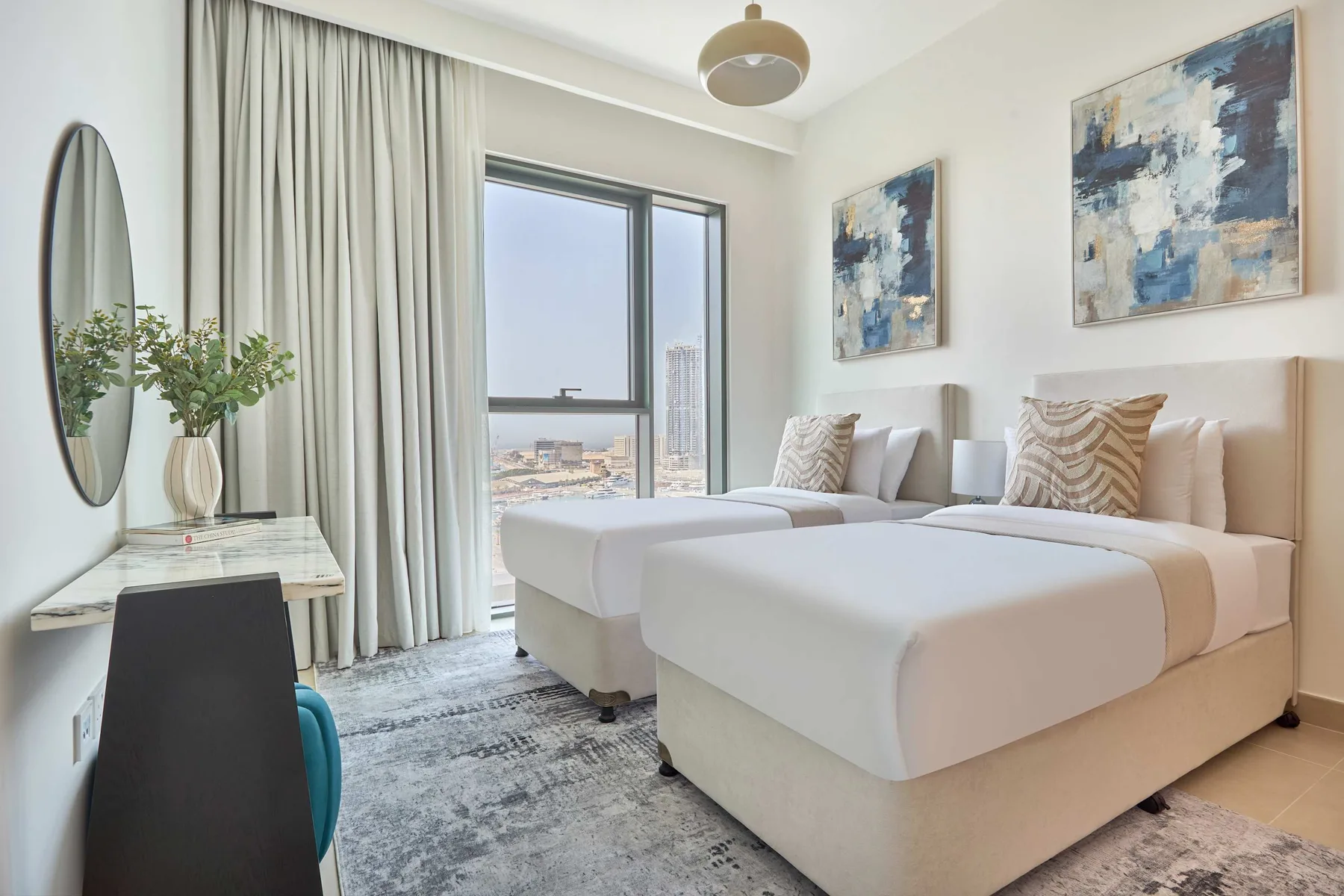 A bright bedroom with two beds, a window, and neutral-toned artwork.