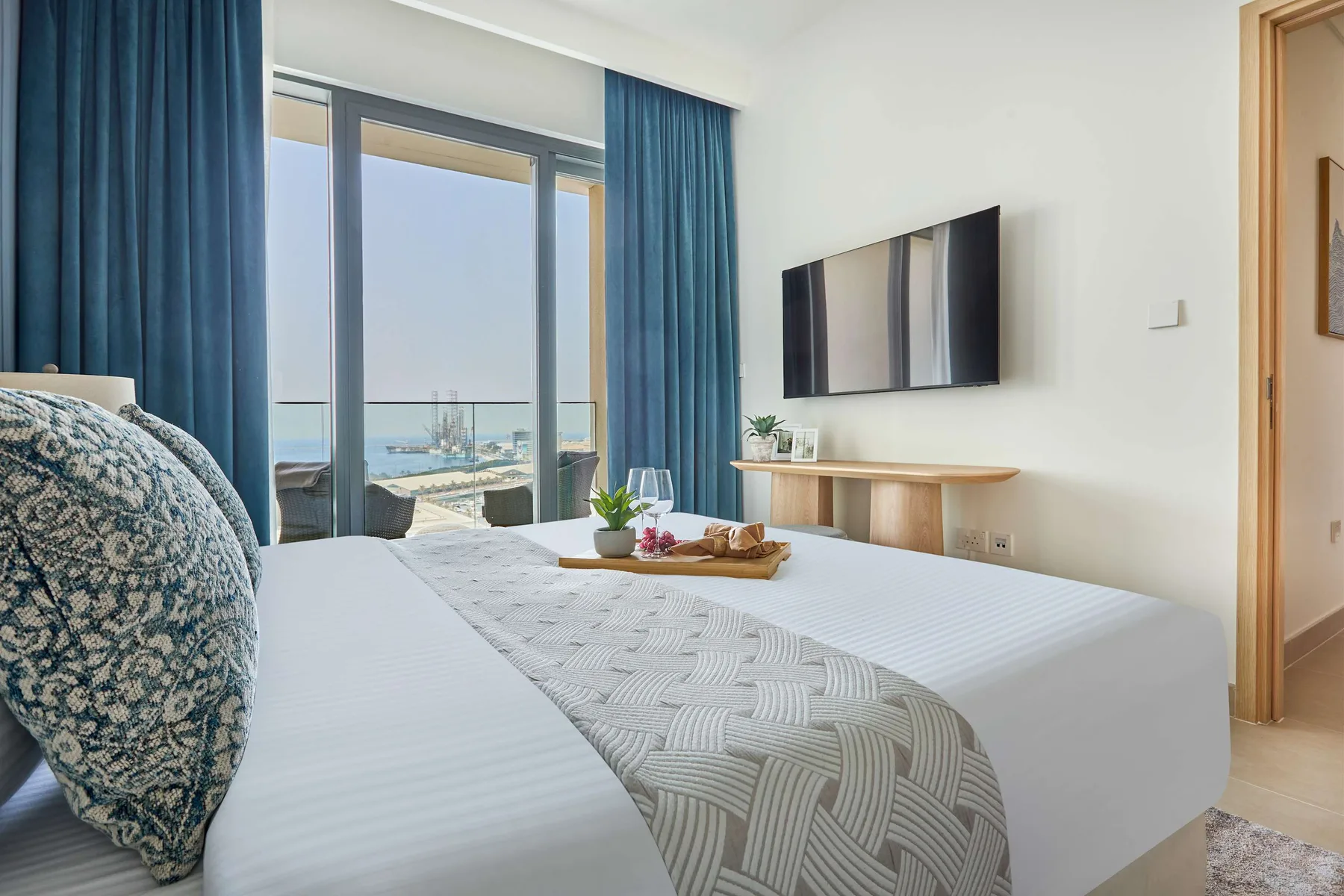 A bright bedroom with a large bed, wall-mounted television, and ocean view through glass doors.