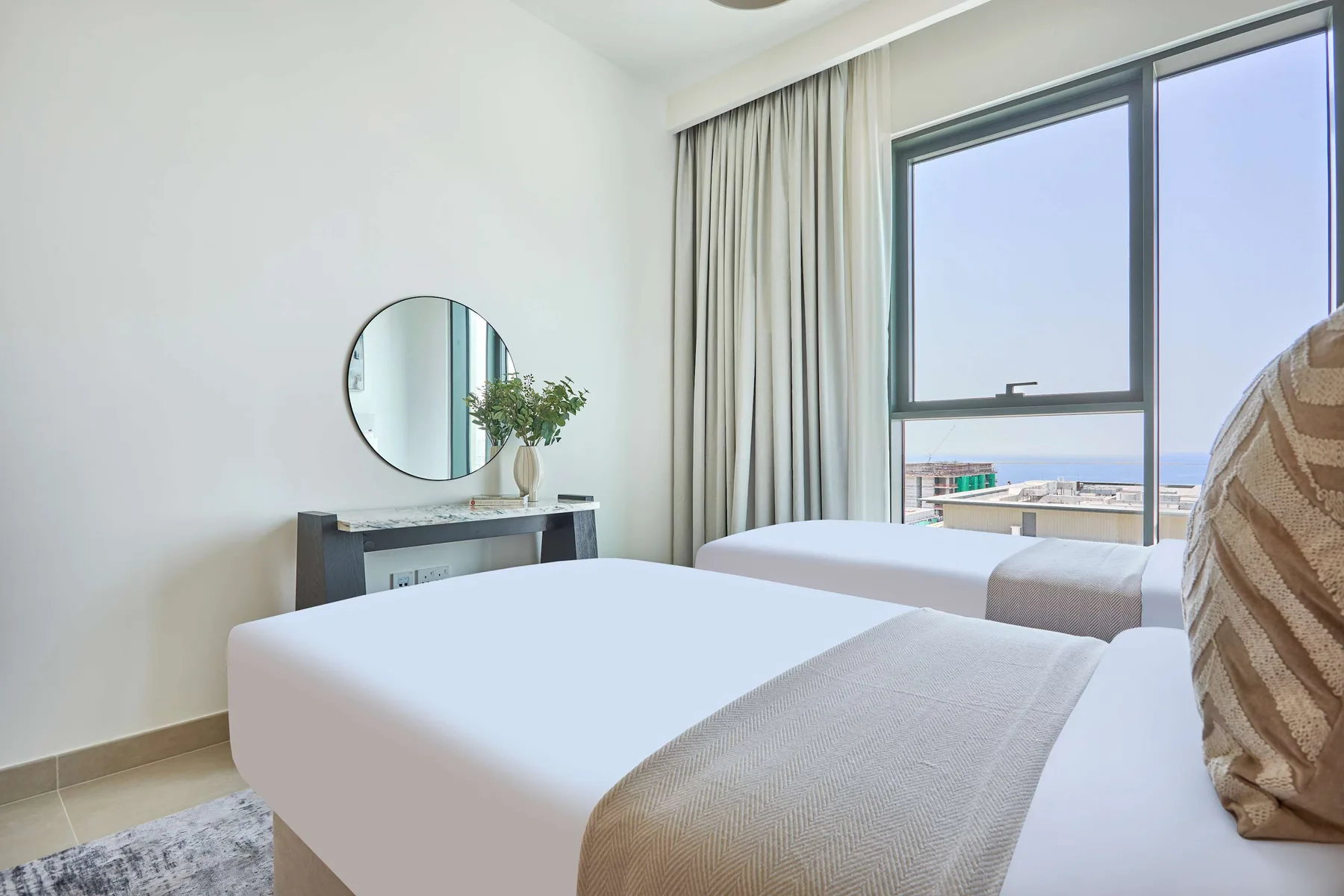 A bright bedroom with two beds, a round mirror, and a large window.