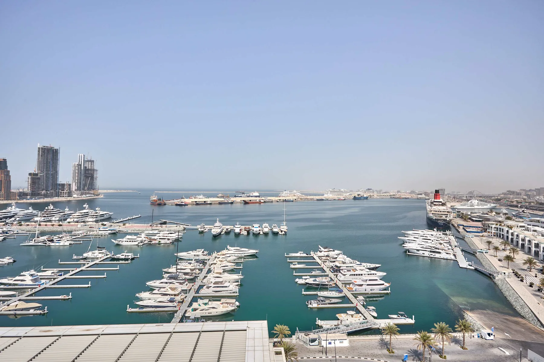 A marina filled with boats stretches along a waterfront promenade beneath a clear sky.