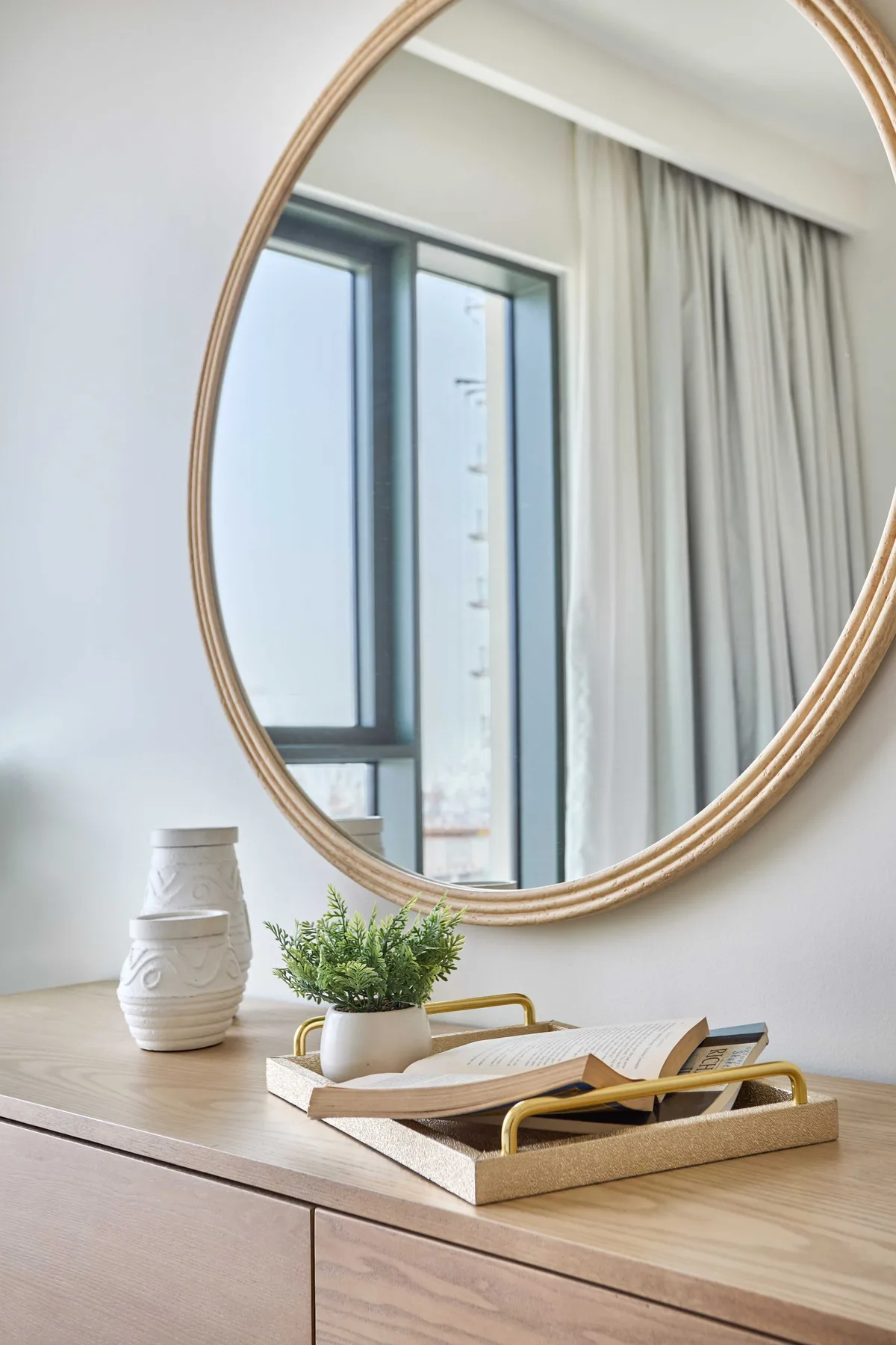 A bedroom dresser holds a decorative tray and plant beneath a large oval mirror.