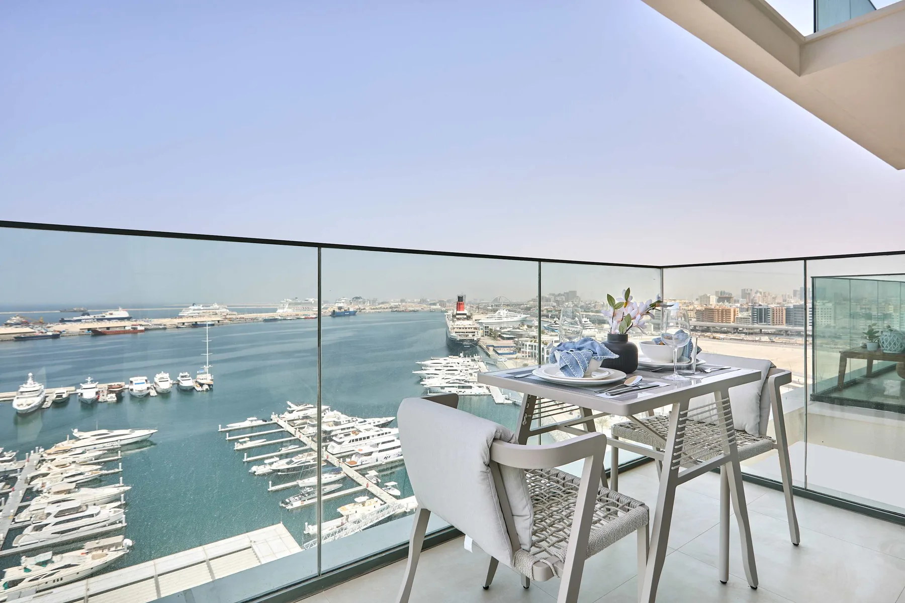 A dining table and seating overlook a marina through glass railings.