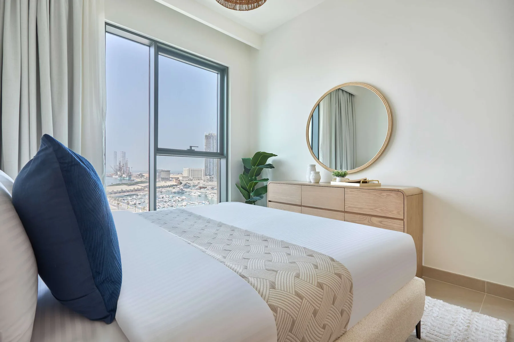 A bright bedroom with a neatly made bed, a round mirror, and a large window.