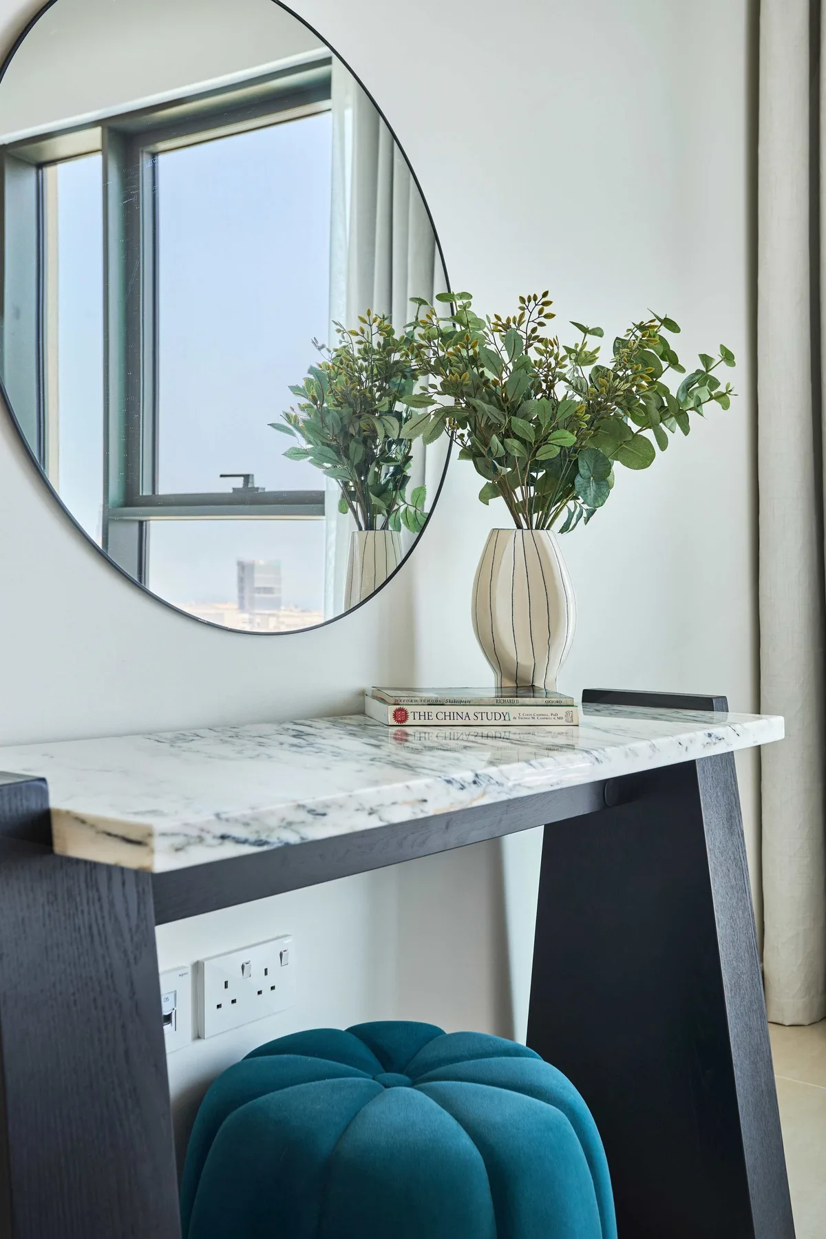 A decorative table stands beneath a round mirror, with flowers in a vase.