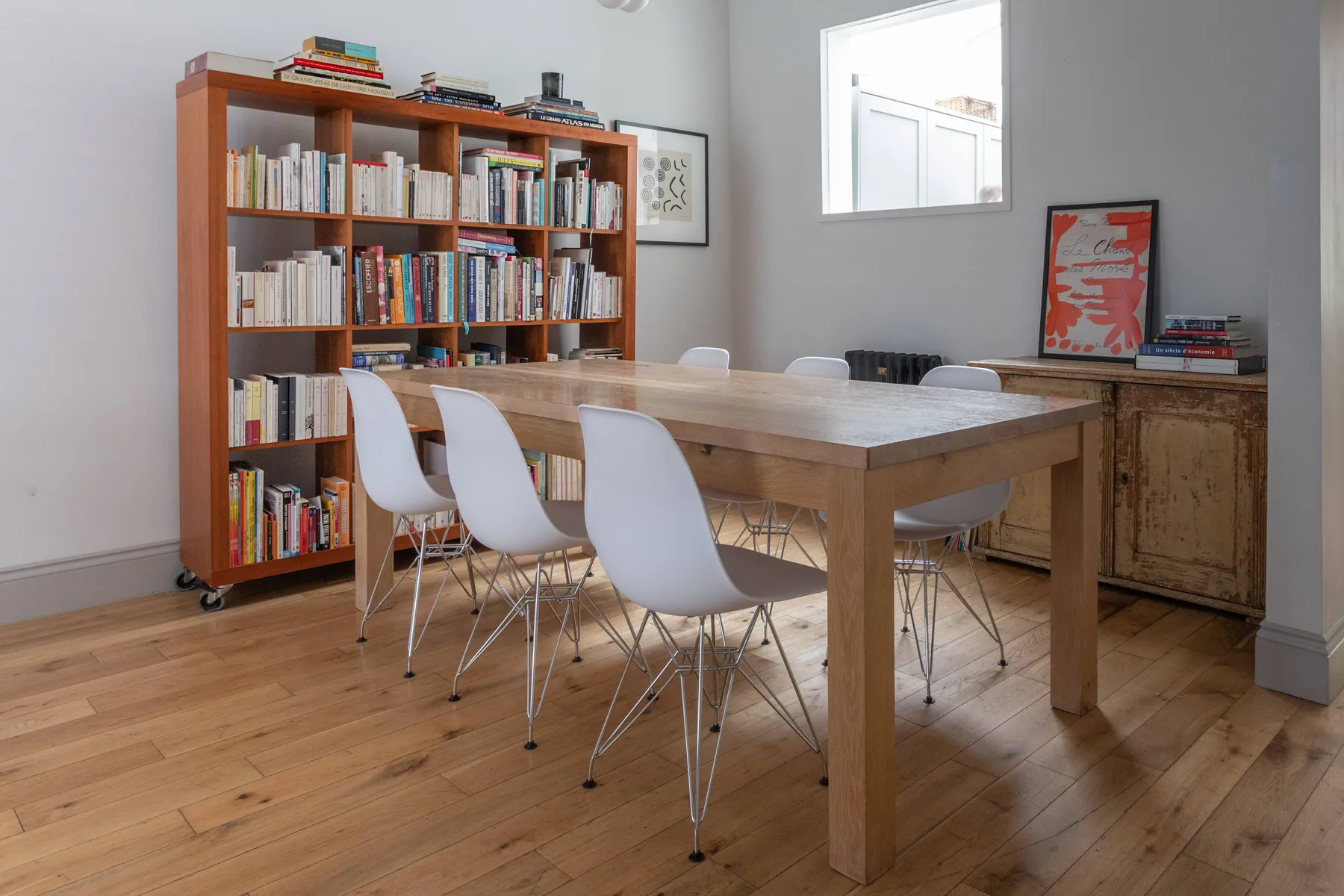 A dining area with a wooden table, white seating, and a bookcase.