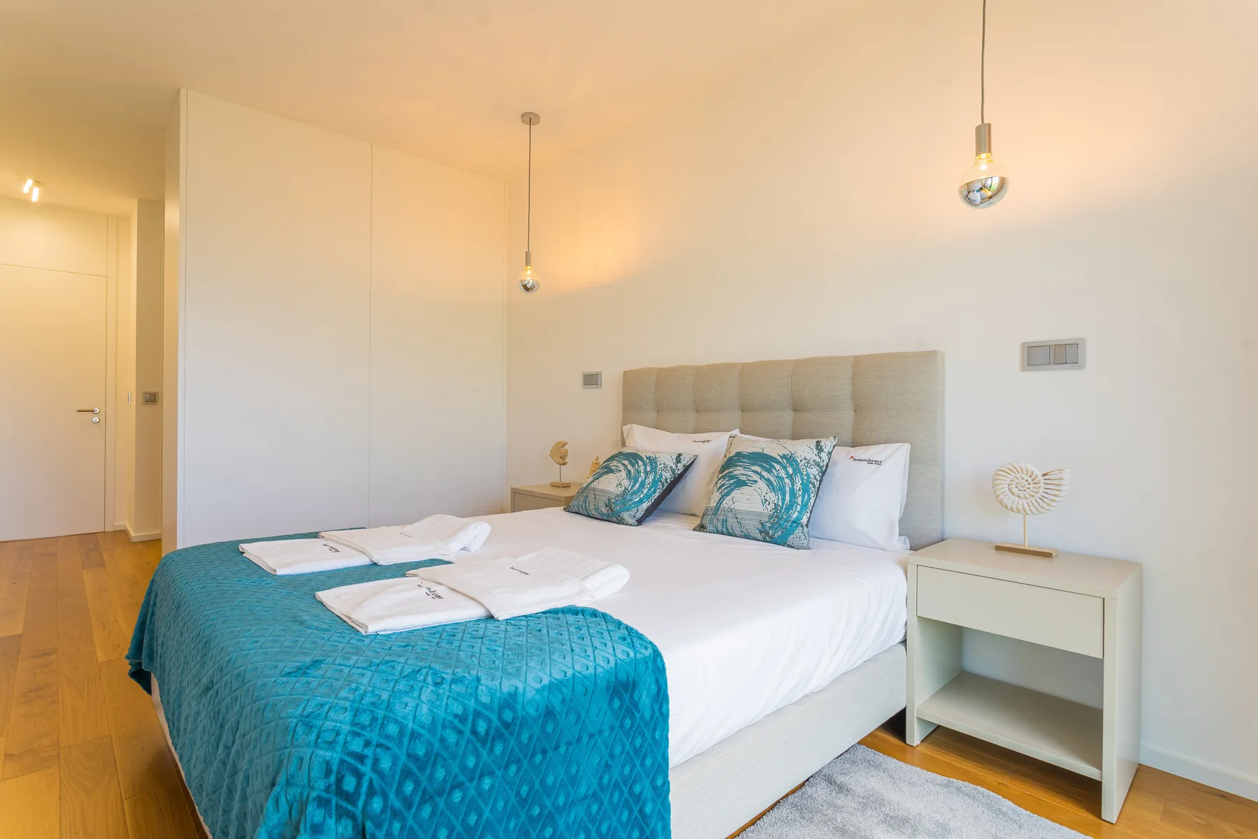 Un dormitorio luminoso con una cama doble vestida con sábanas blancas y una colcha turquesa.