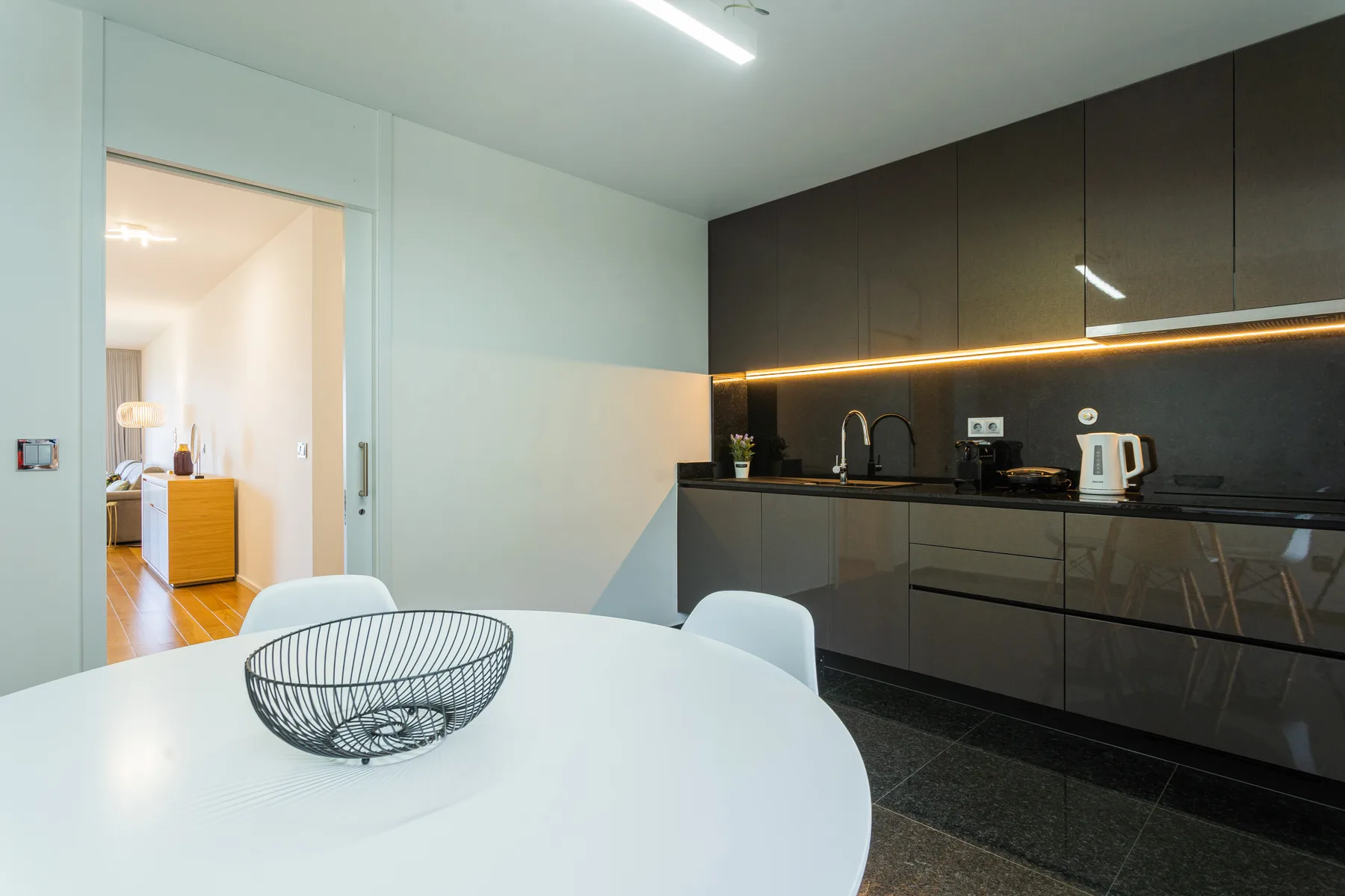 A bright dining area with a white table beside a dark modern kitchen.