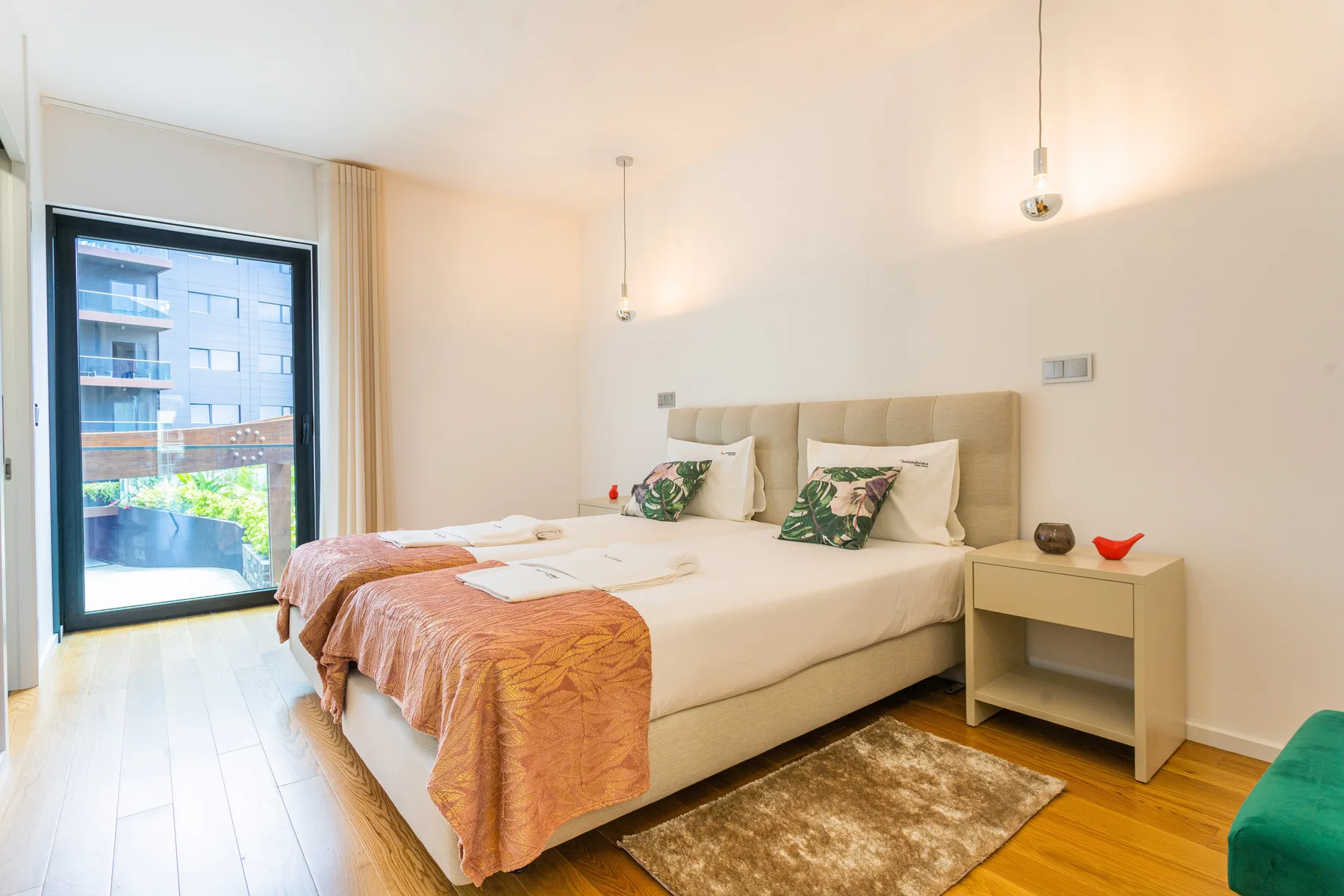 Un dormitorio luminoso con una cama grande junto a una ventana con vistas a edificios cercanos.