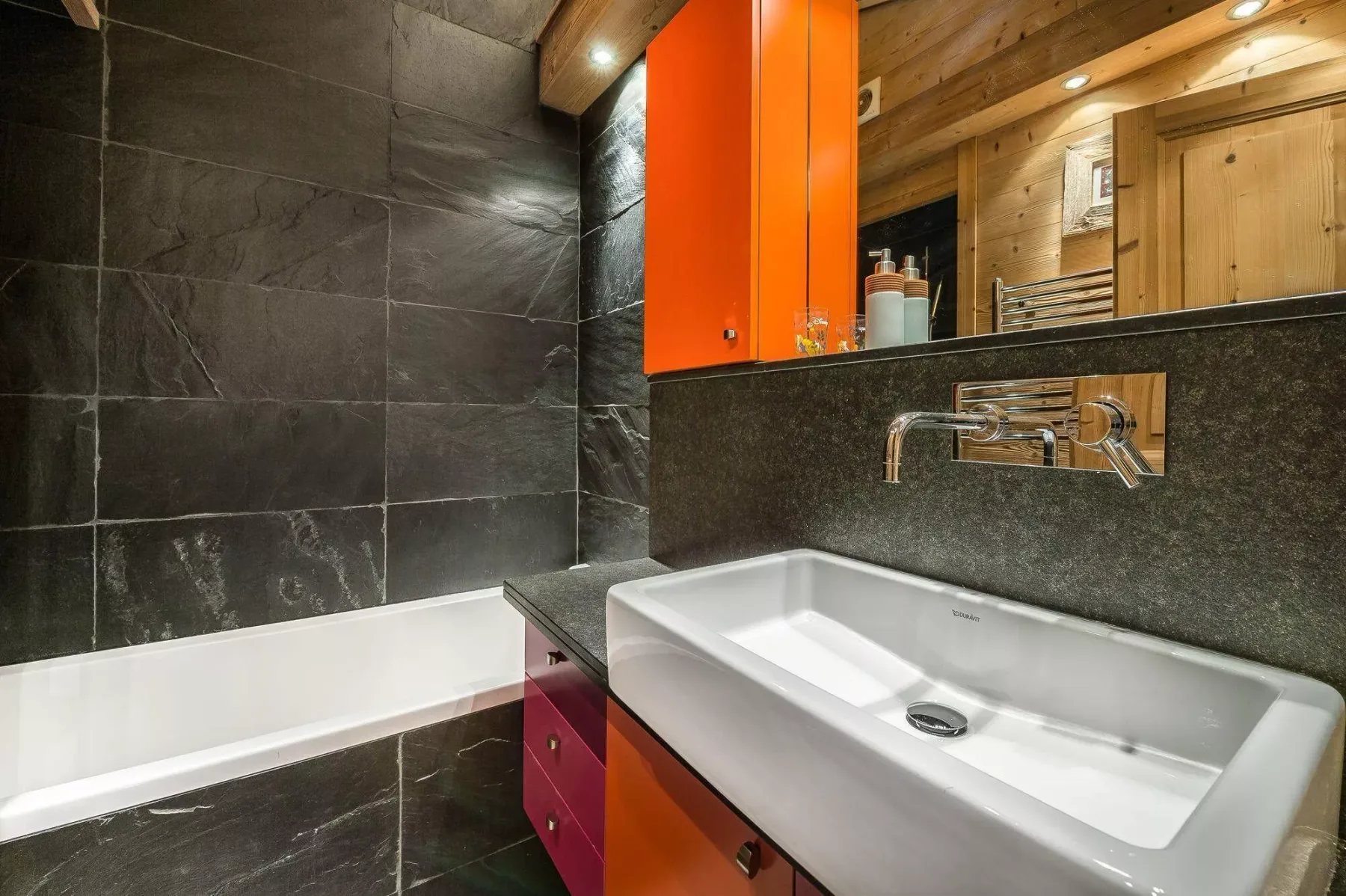Un baño con bañera blanca y lavabo blanco junto a azulejos oscuros.