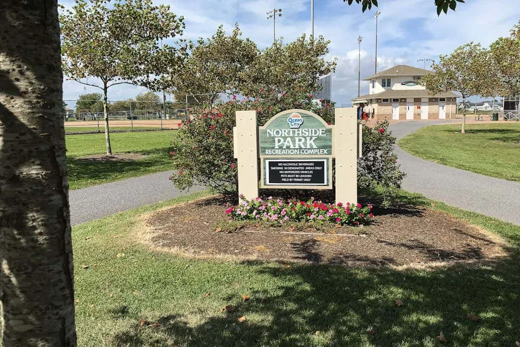 A park entrance sign stands beside a paved path and landscaped flower bed.