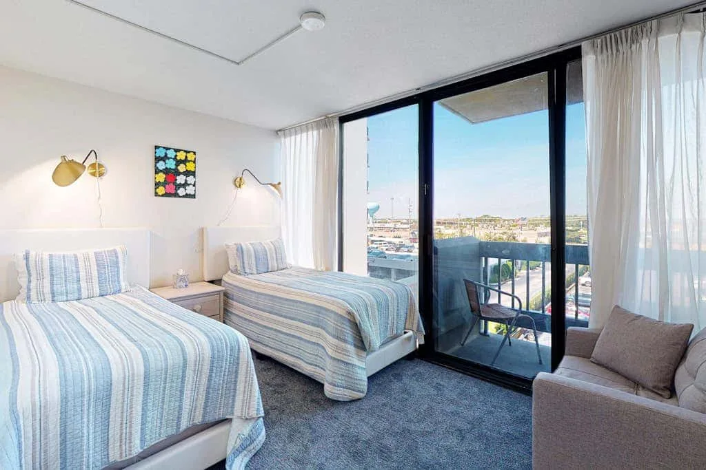 A bright bedroom with two beds beside large glass doors opening onto a balcony.