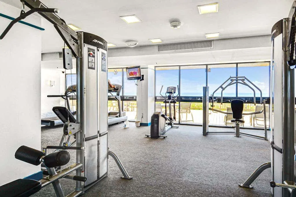 A bright gym with exercise machines and large windows overlooking the water.
