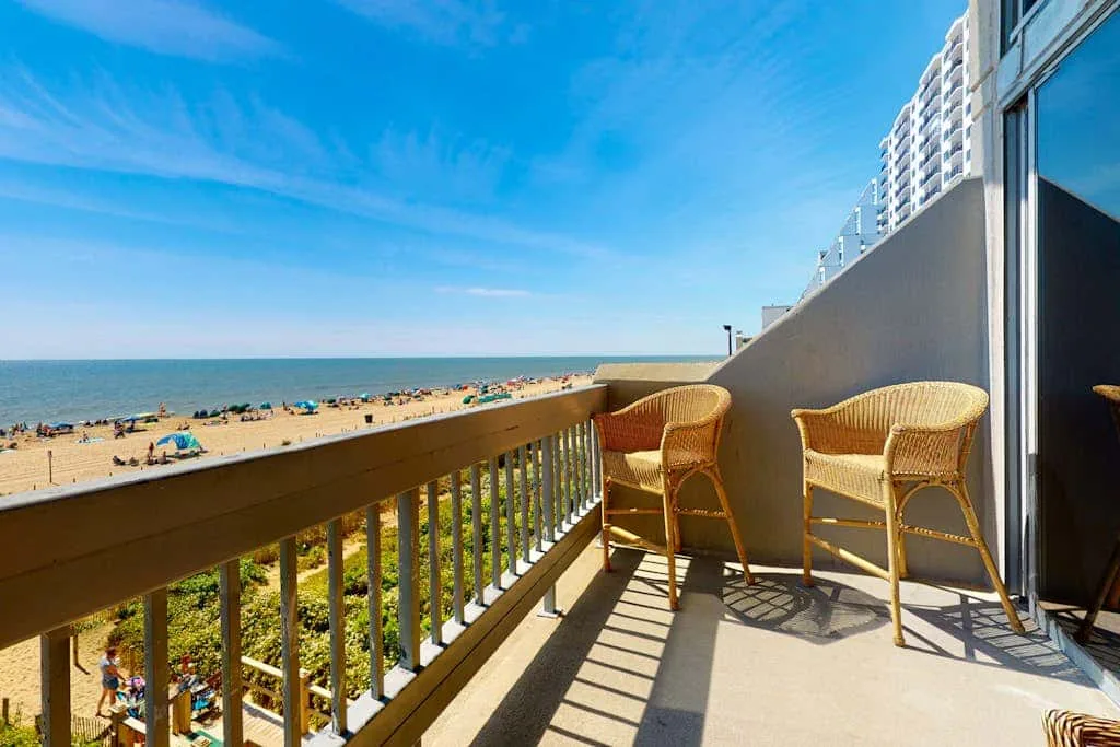 A sunny balcony overlooks a sandy beach and the sea, with wicker seating.