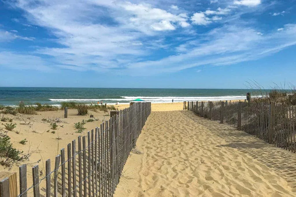 A sandy path through beach fencing leads toward the ocean under a cloudy sky.