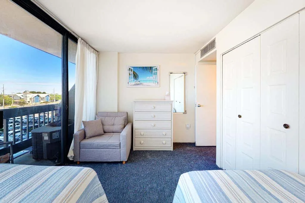 A bright bedroom with two beds, a dresser, and sliding glass doors opening onto a balcony.