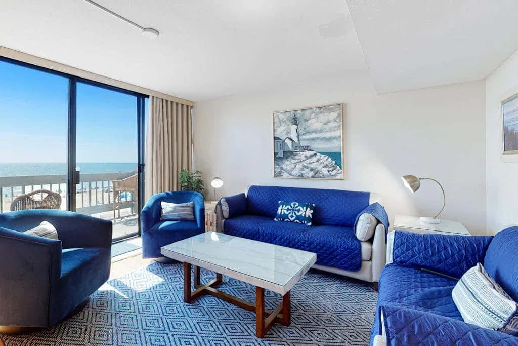 A bright living room with blue seating, a glass coffee table, and a balcony view.