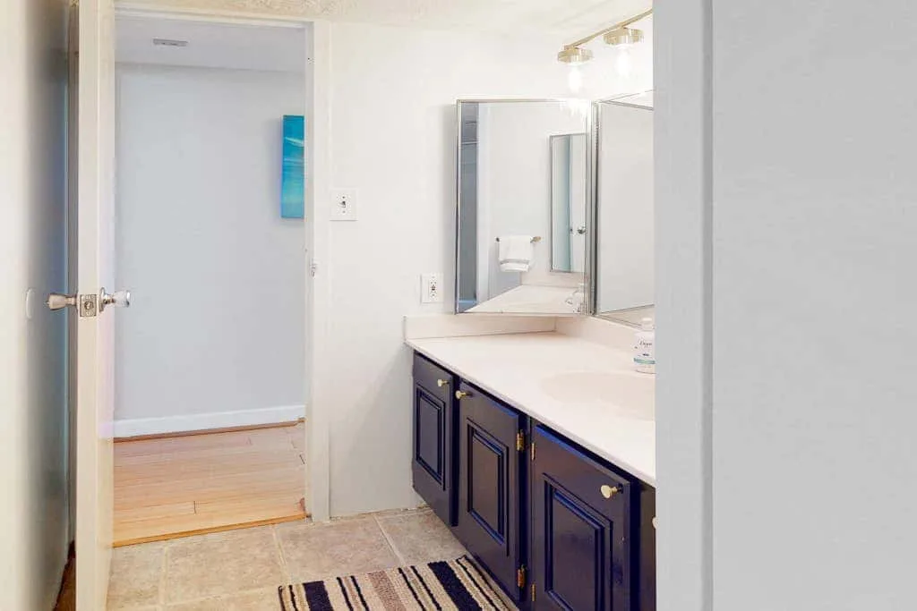 A bathroom vanity with mirrors and a striped rug sits beside an open doorway.