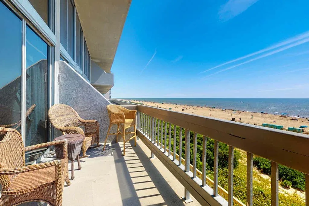 A balcony with wicker seating overlooks a sandy beach and the ocean.