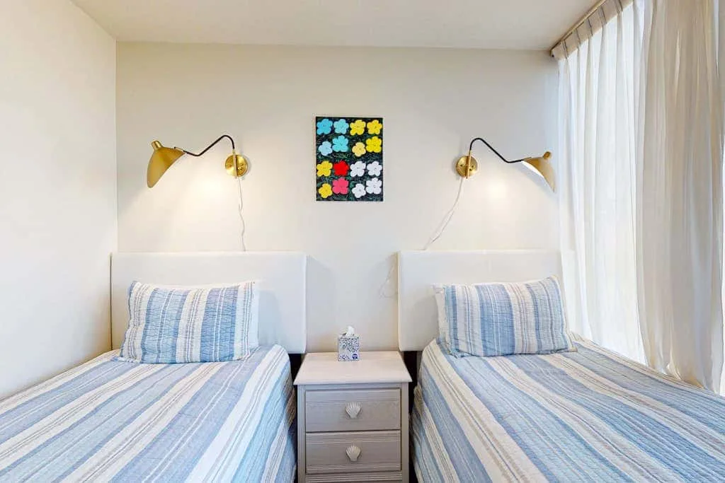 A bright bedroom with two beds, striped bedding, and a small bedside table.