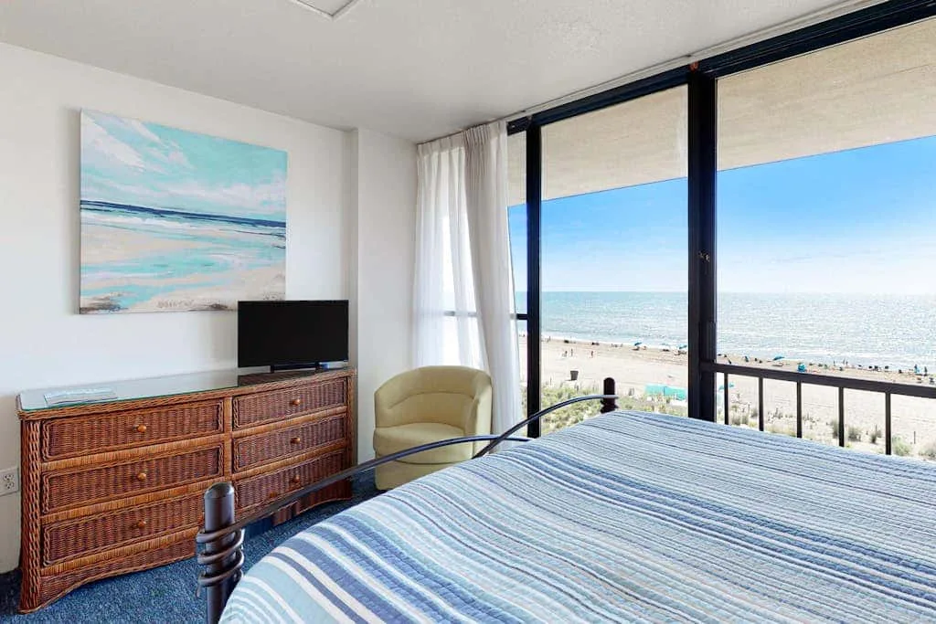 A bedroom with a striped bedspread and large windows overlooking the beach.