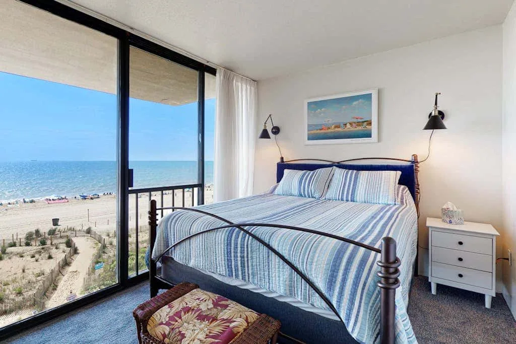 A bedroom with a striped bed beside floor-to-ceiling windows overlooking the beach.