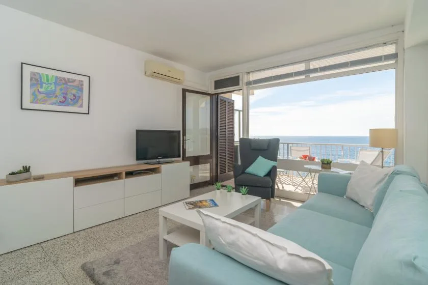 A bright living room with seating, a television, and a balcony overlooking the sea.
