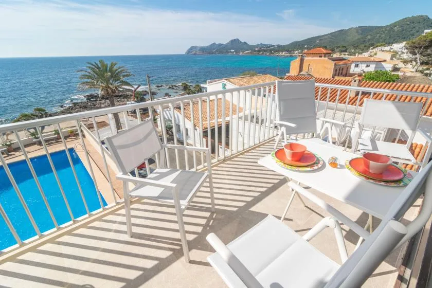 A sunny seaside balcony has a dining table, outdoor seating, and views of the sea.