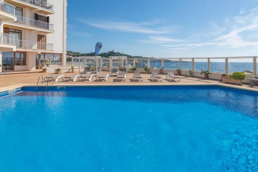 A swimming pool beside a sunlit terrace with seating and a sea view.