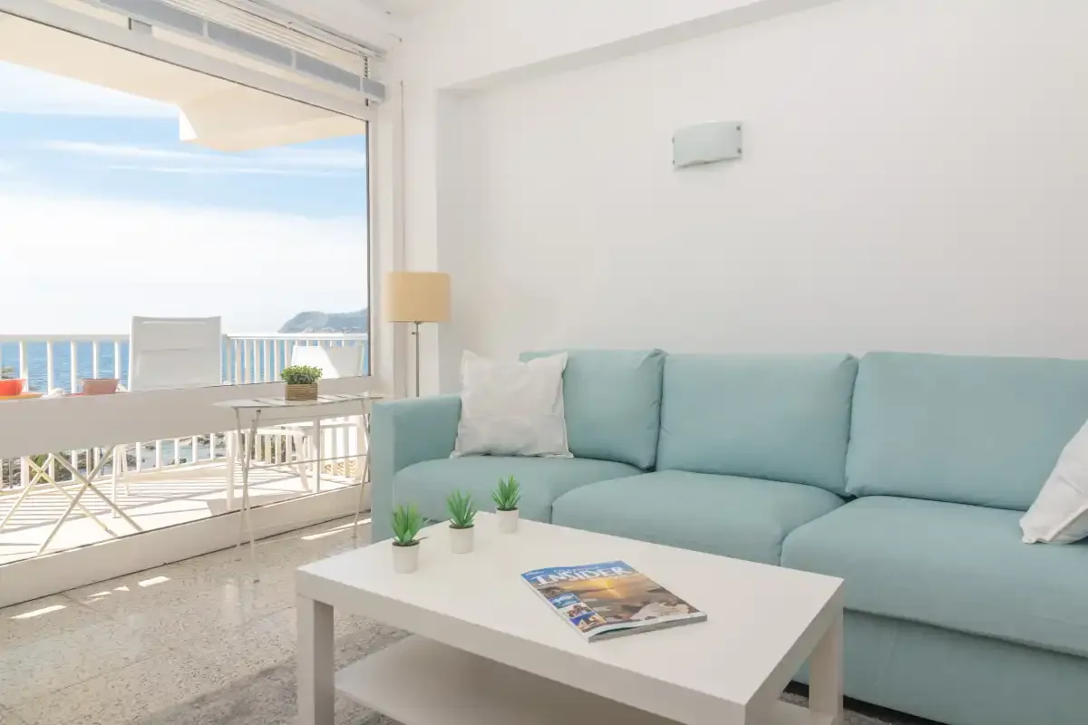 A bright living room with pale blue seating beside a large window overlooking the sea.
