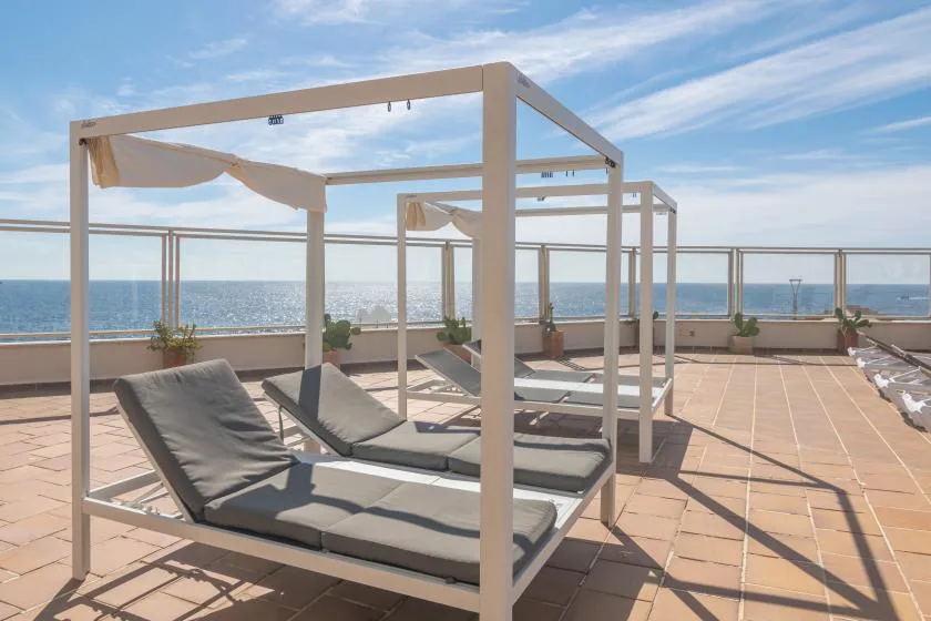 A seaside terrace with cushioned loungers beneath open-frame canopies.