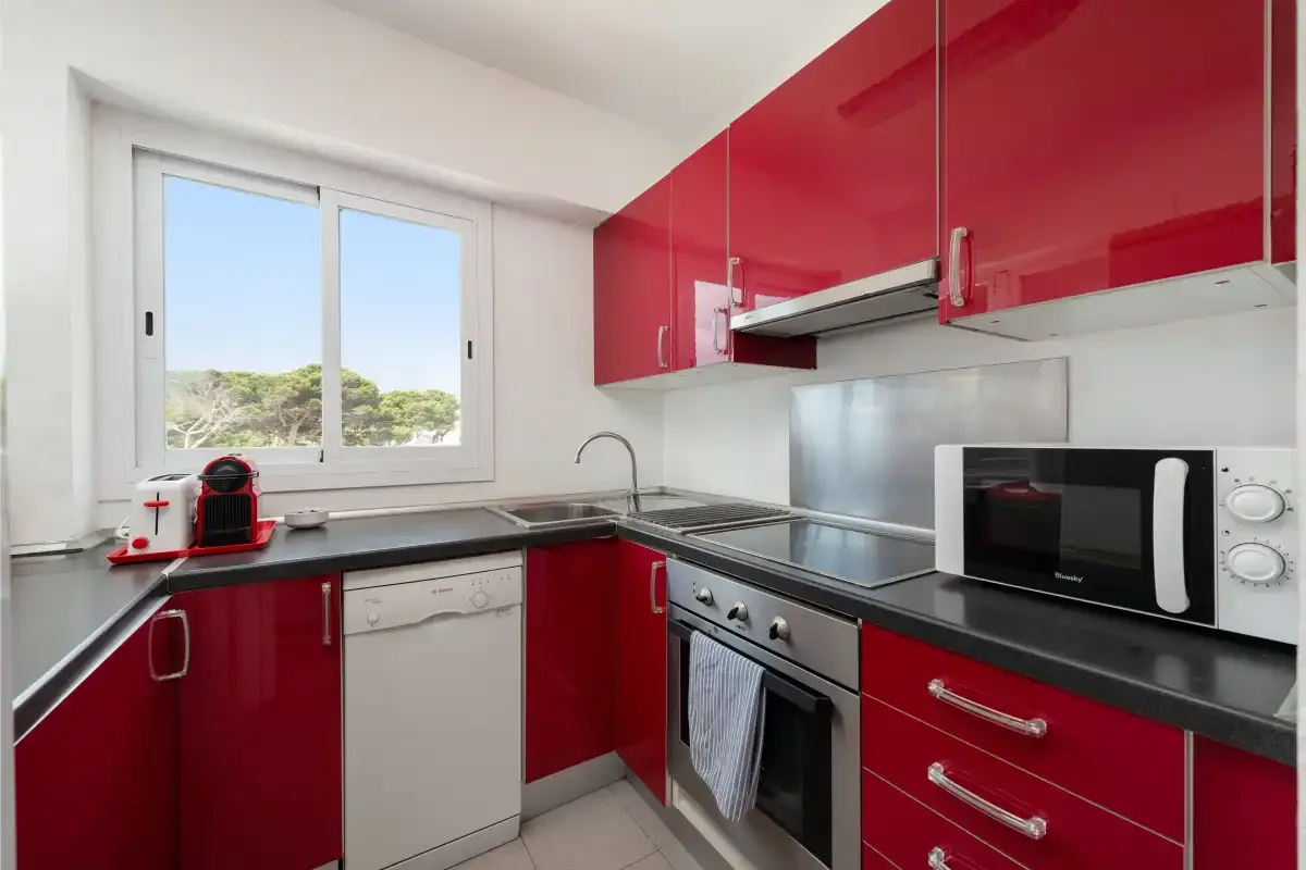 A kitchen with red cabinets, dark countertops, and a window.