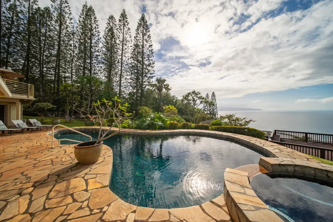 A curved outdoor swimming pool beside a stone patio, with trees and ocean beyond.