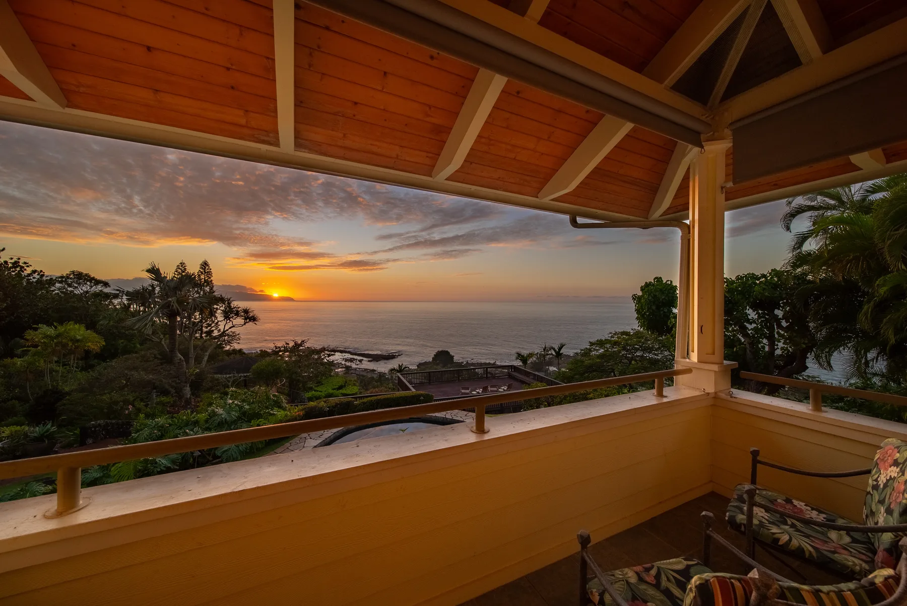 A covered veranda overlooks the ocean at sunset, with seating beside the railing.