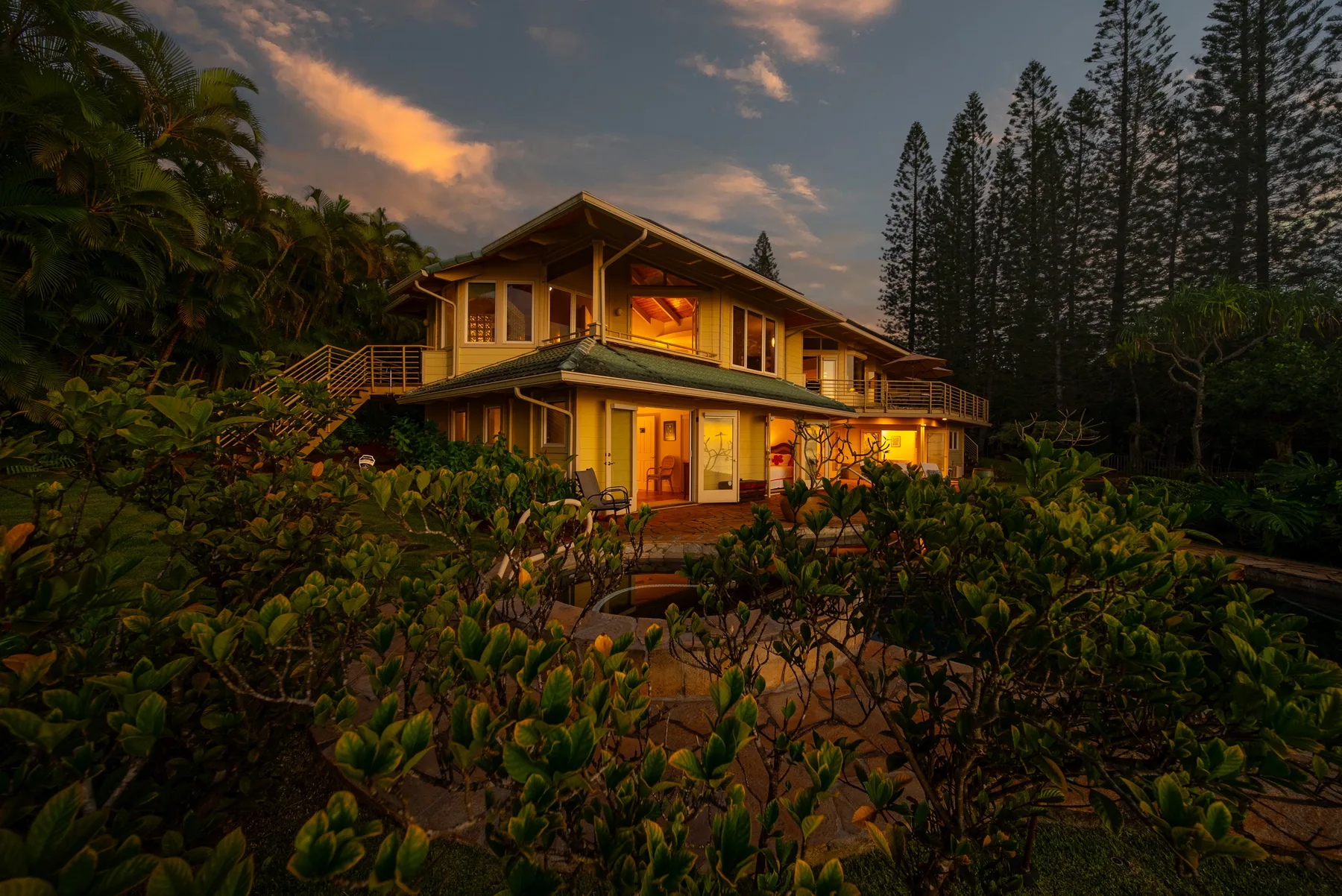 A warmly lit house stands amid dense greenery at dusk.