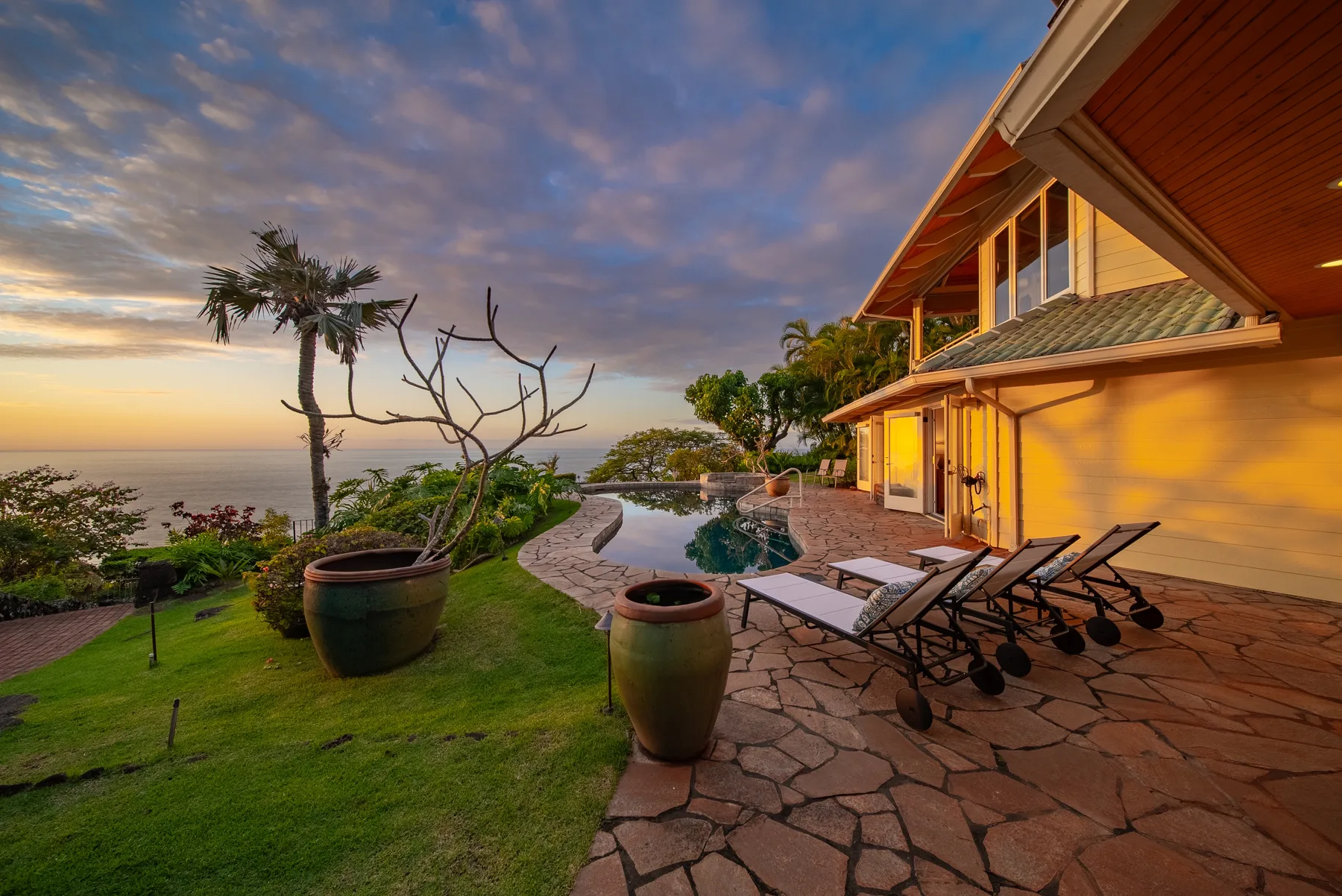 A landscaped outdoor pool and patio beside a house overlook the ocean at sunset.