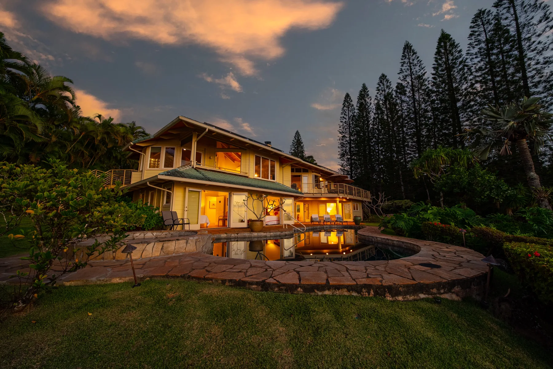 A large house is lit at dusk beside a curved pool and landscaped garden.