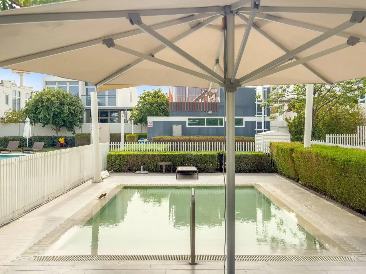 Una piscina está bajo un toldo grande, con setos y edificios cerca.