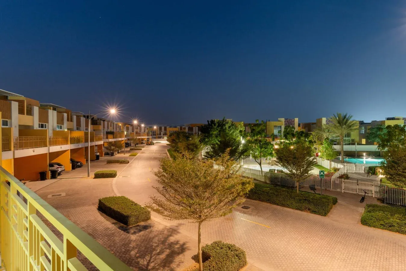 Patio de un conjunto residencial al anochecer, con senderos, vegetación y edificios iluminados.