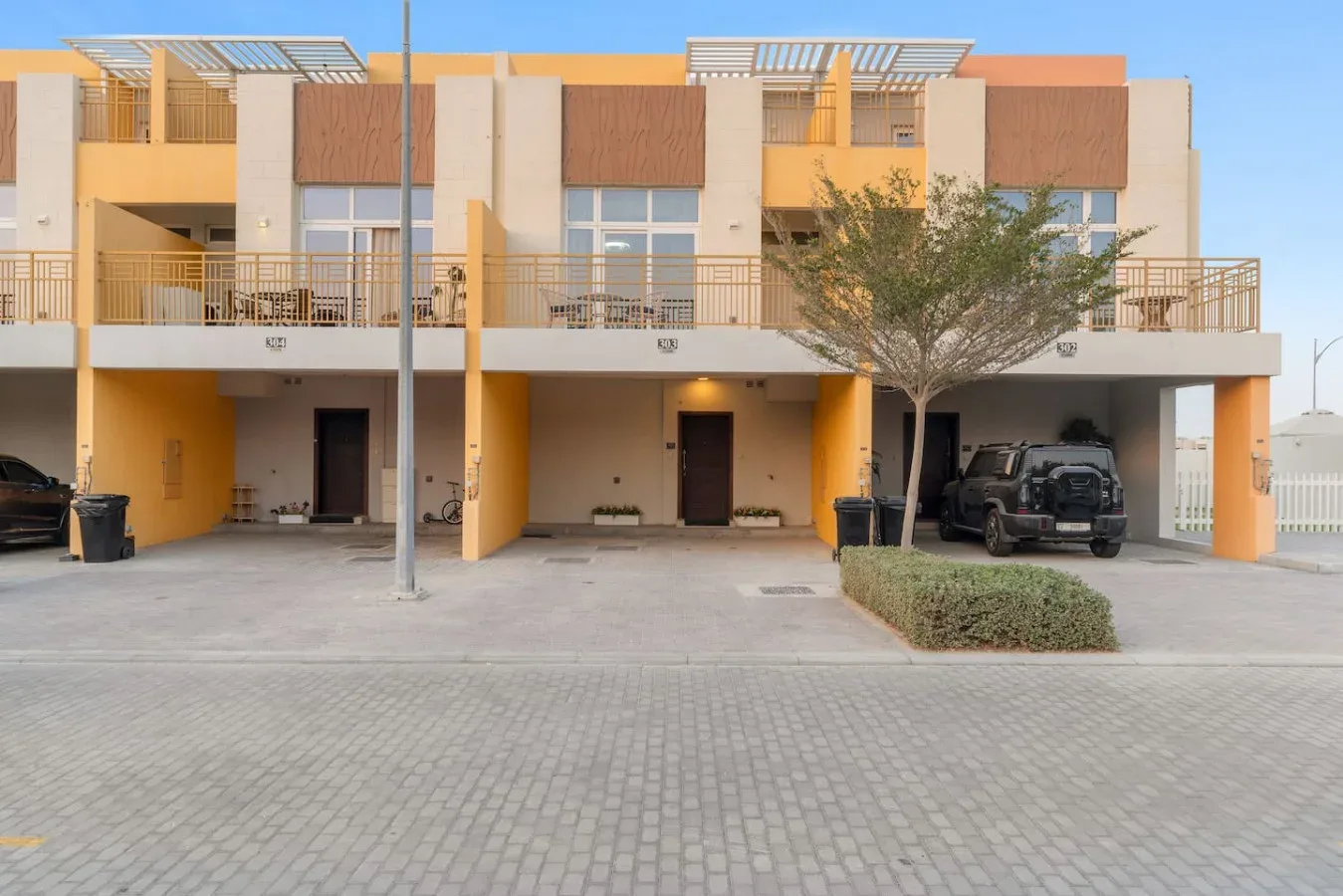 Una hilera de casas adosadas modernas da a una zona de estacionamiento pavimentada, con un árbol delante.
