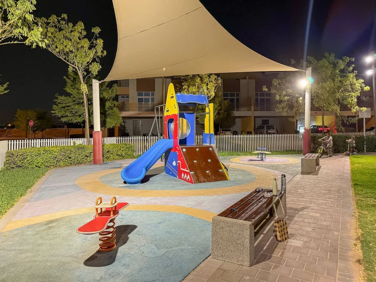 Un parque infantil de noche con juegos de colores bajo una gran cubierta de sombra.