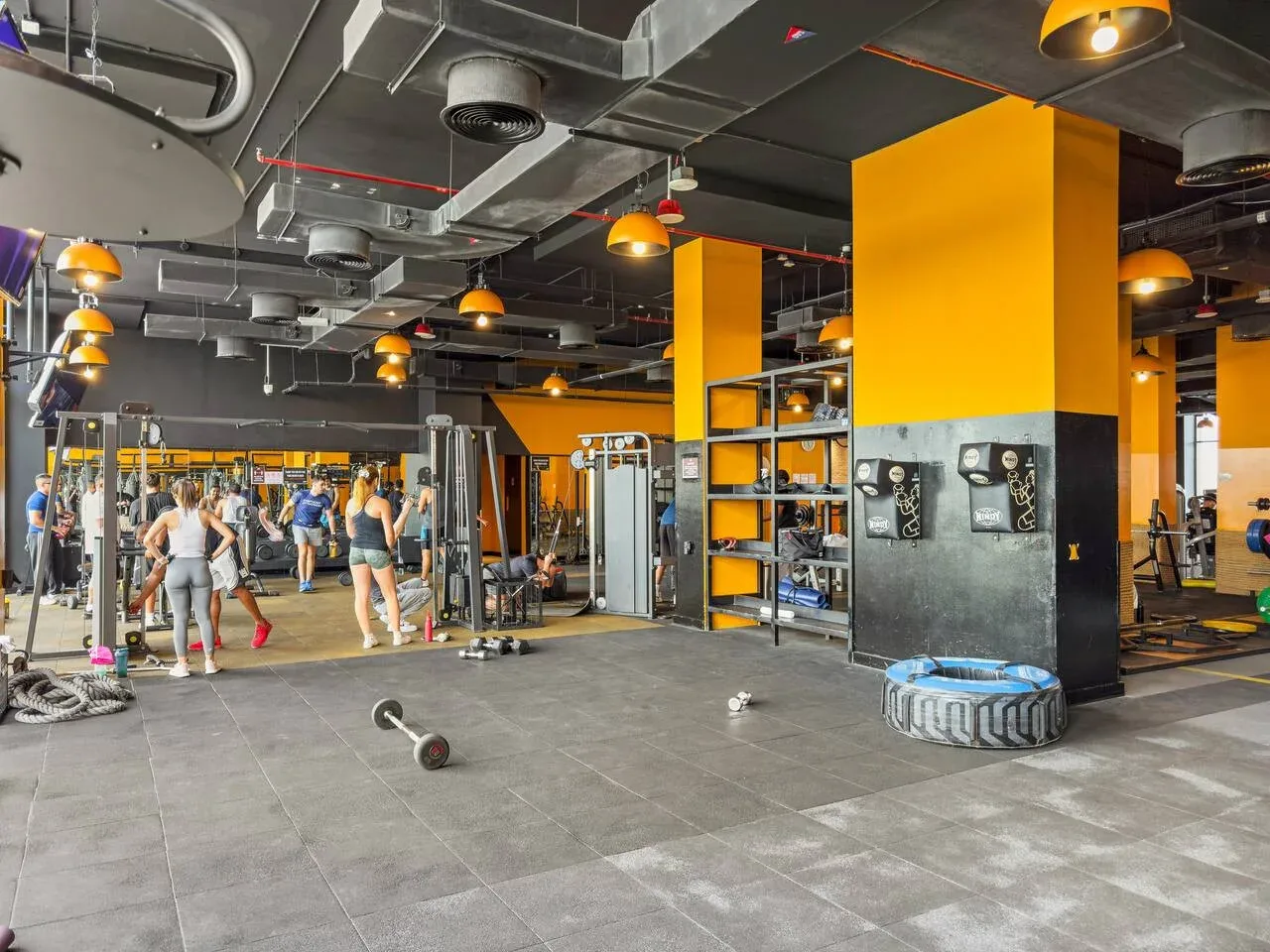 Una zona de gimnasio con equipo de ejercicio y personas entrenando bajo conductos expuestos.