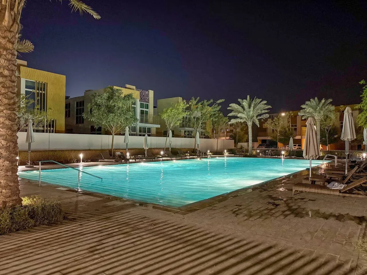 Una piscina al aire libre iluminada por la noche, rodeada de palmeras y asientos.