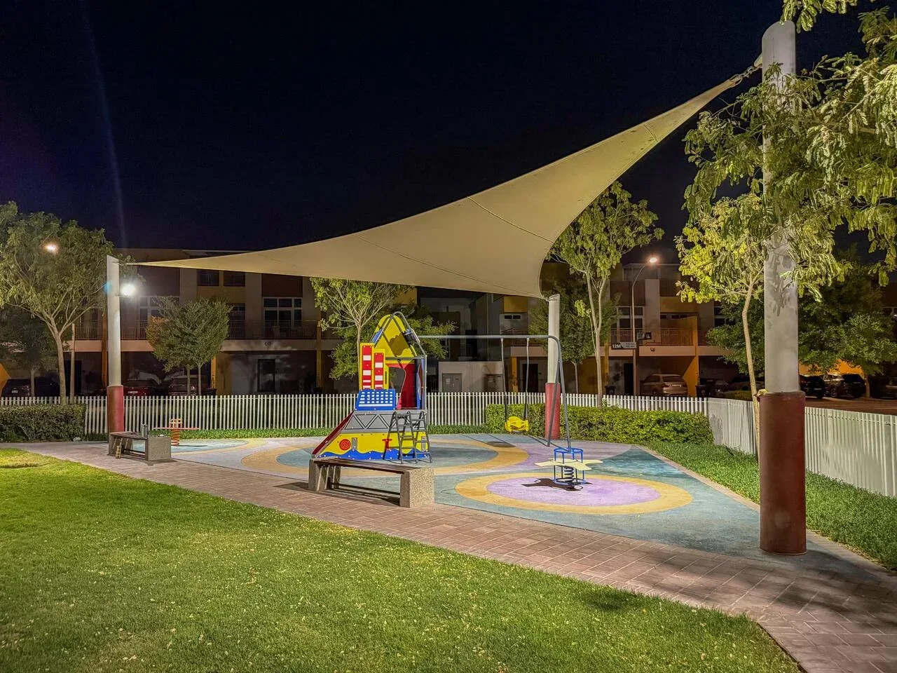Un parque infantil de noche bajo una gran cubierta de sombra, con juegos coloridos y césped alrededor.