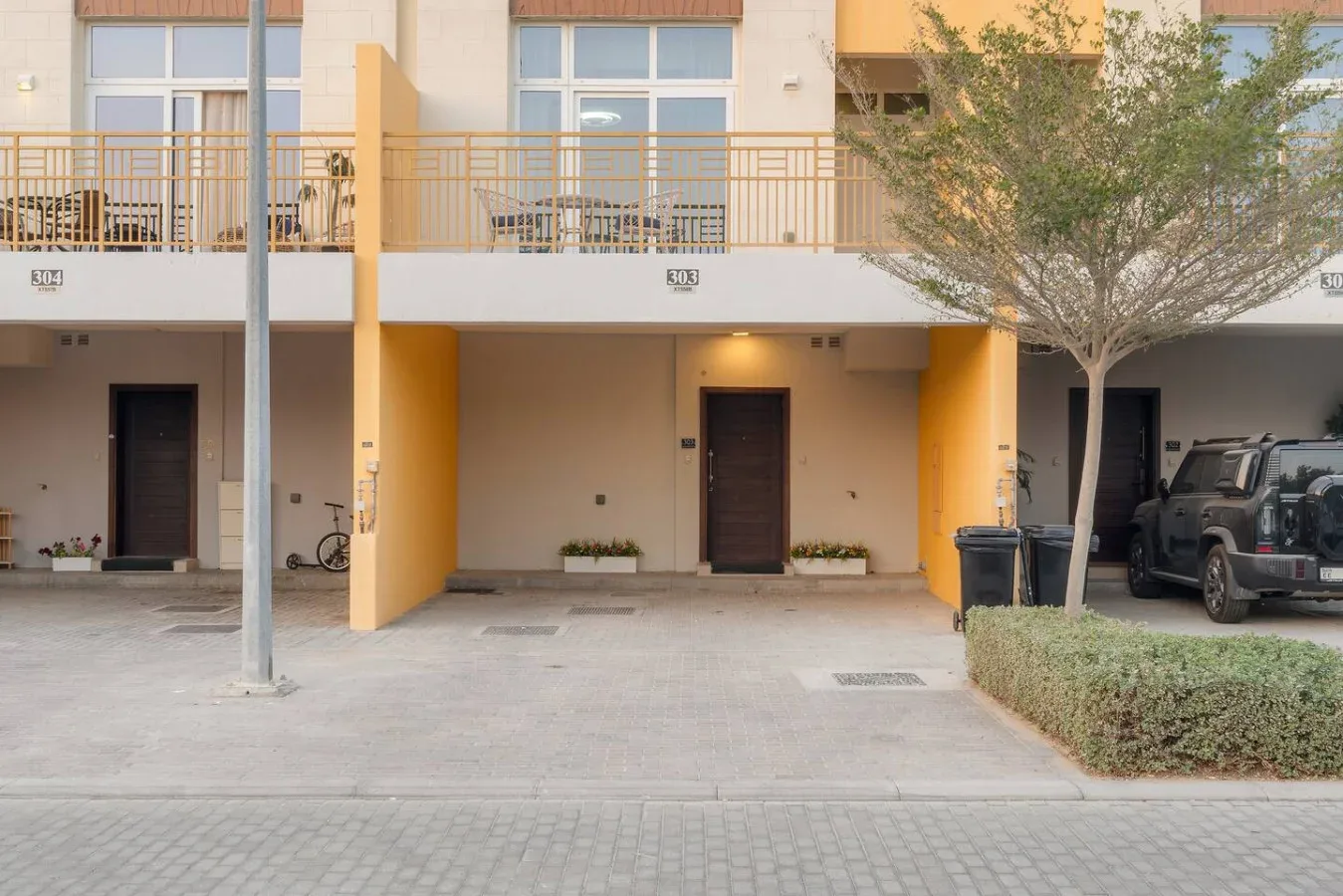 Entradas en planta baja bajo balcones, con una zona pavimentada y vehículos estacionados.