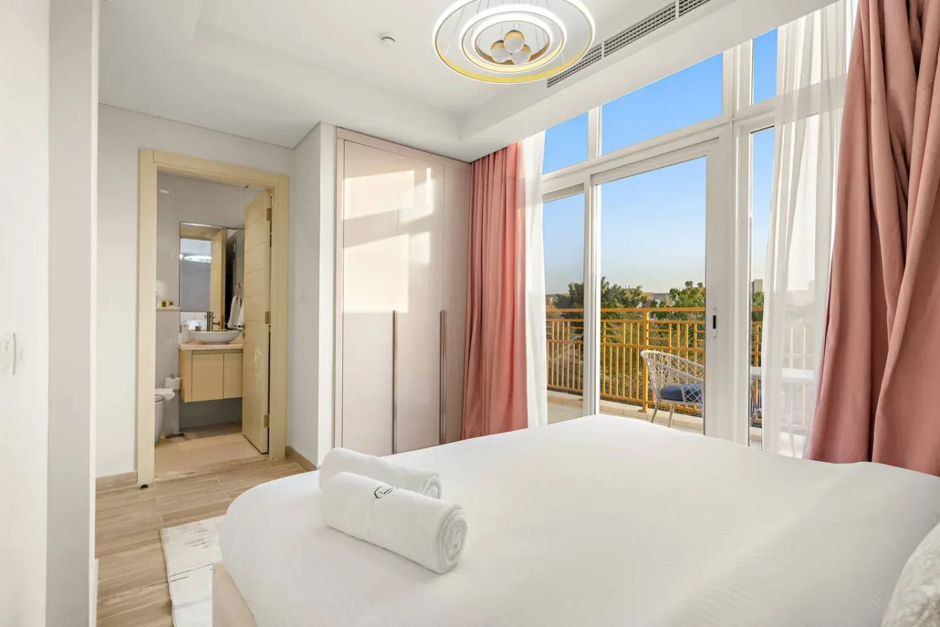 Un dormitorio luminoso tiene una cama bien tendida junto a grandes ventanas y cortinas rosas.