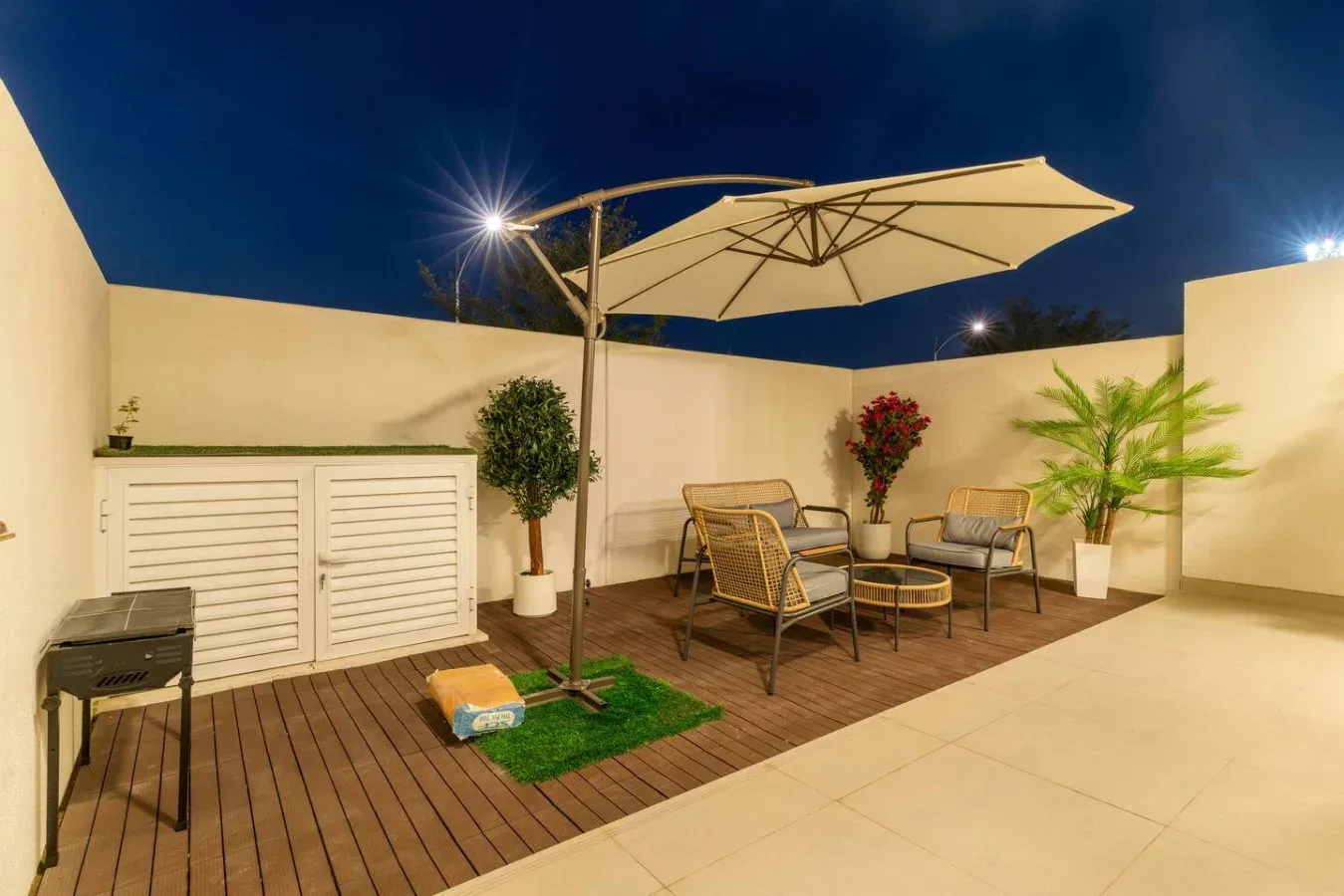 Un patio exterior nocturno tiene una tarima de madera, asientos y una sombrilla grande.