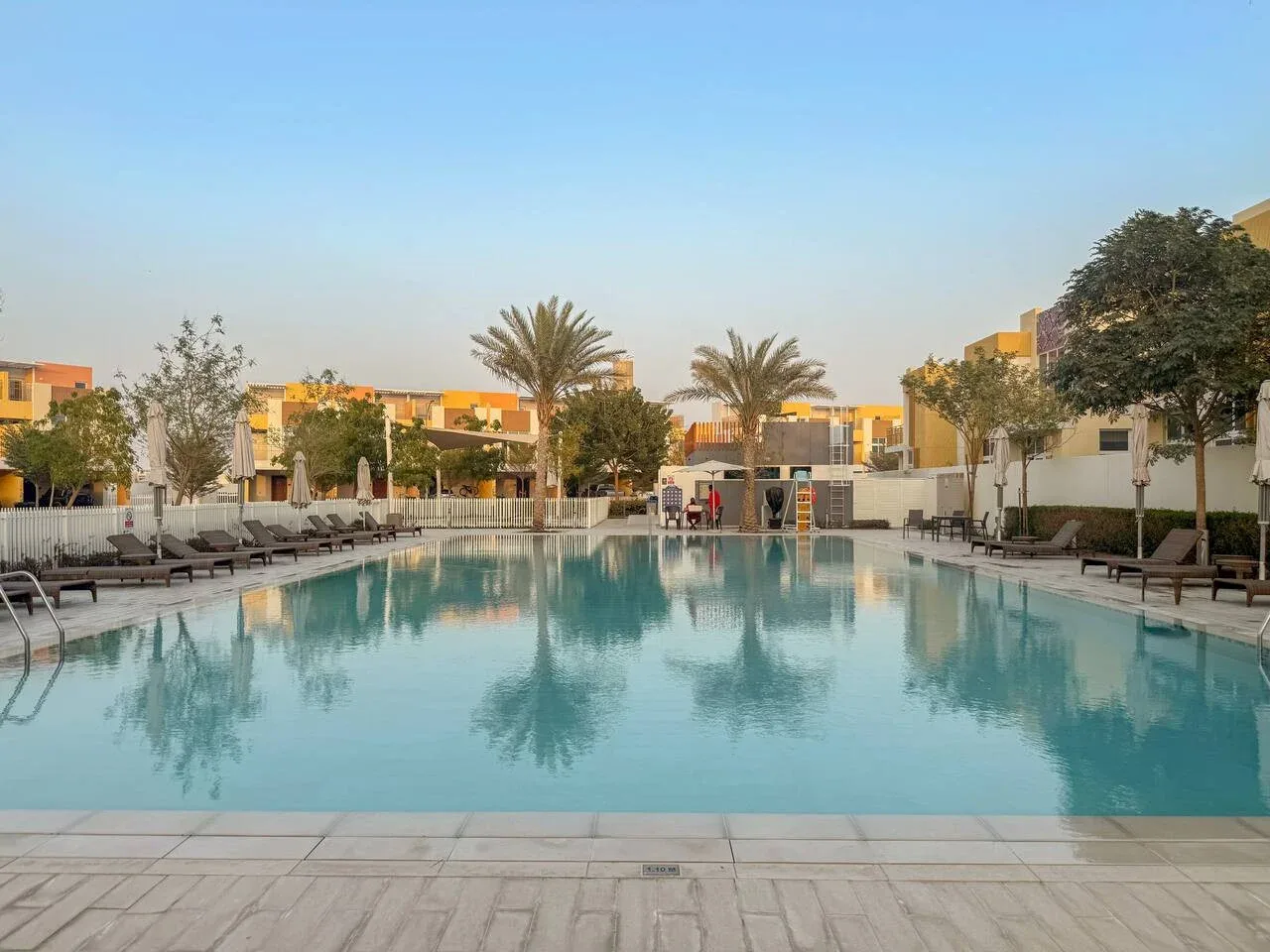 Una piscina al aire libre está rodeada de asientos, árboles y edificios bajos.
