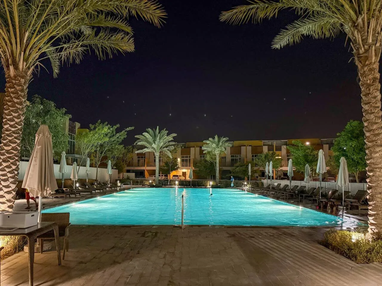 Una piscina iluminada por la noche, enmarcada por palmeras y asientos al aire libre.