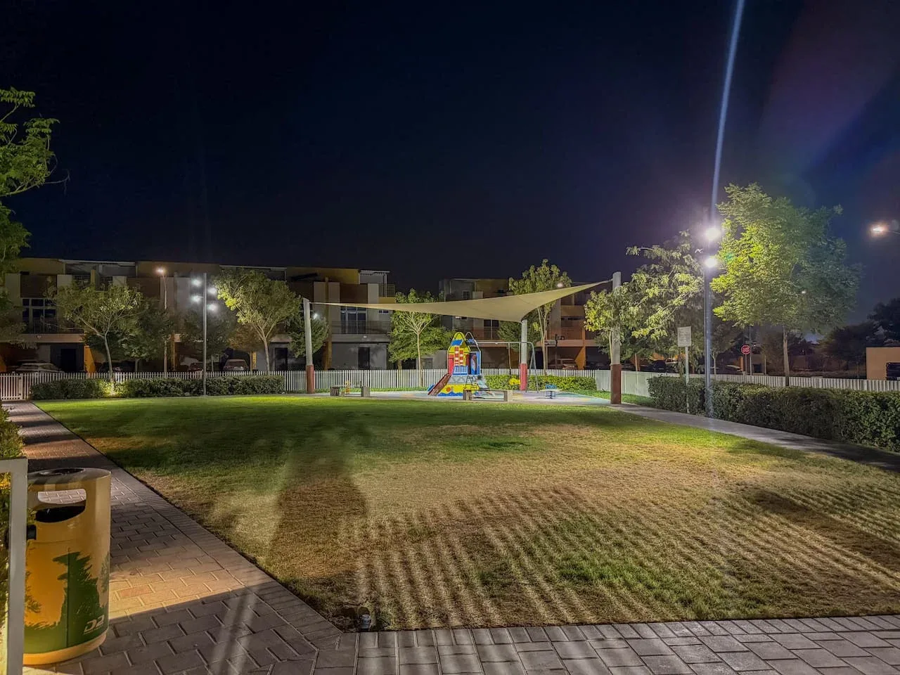 Un patio ajardinado al aire libre por la noche, con césped, juegos infantiles y edificios alrededor.